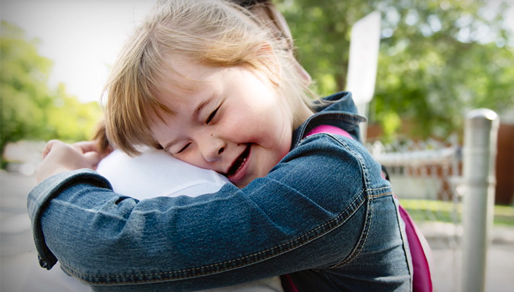 A child with brown hair and Down syndrome hugs an adult tightly while smiling, outside.