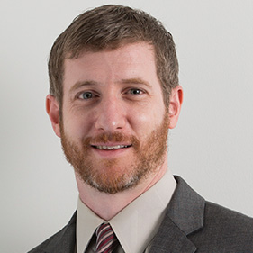 A man with brown hair wearing a gray suit, cream-colored shirt and burgundy tie smiles for a portrait