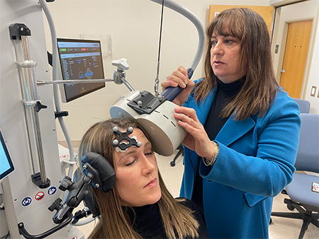 A woman with a black tracker on her forehead sits with her eyes closed while another woman maneuvers a beige magnetic coil 