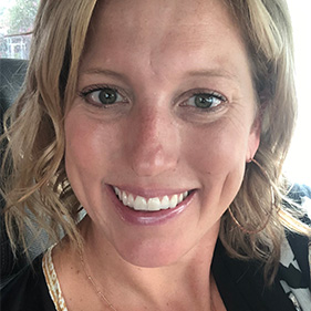 A woman with medium-length blonde hair, wearing a black shirt, gold necklace and gold earrings smiles from inside a car. 