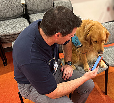 A man sits on a bench bending down to hold a cell phone to the face of a golden retriever wearing a blue service dog vest 