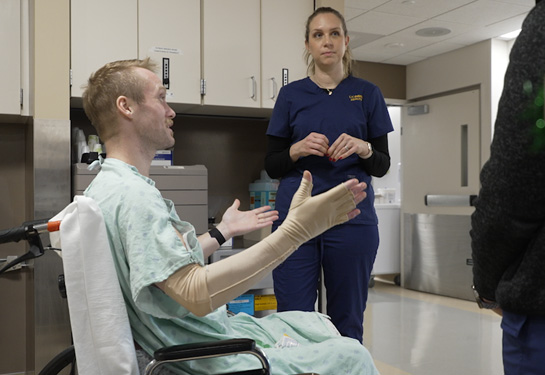 Man on wheelchair motions with his bandaged right arm while talking with burn center nurses.