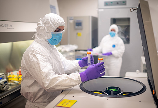 Person wearing blue mask and white PPE suit places vile in machine near another person in a blue mask and white PPE suit 