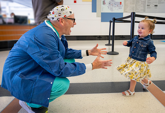 Man in nurse uniform holds arms out and toddler girl with pigtails runs toward him smiling.