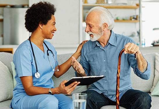 A woman in blue scrubs and holding a tablet sits on a couch next to an older, white-haired man resting is hand on a wooden cane. 