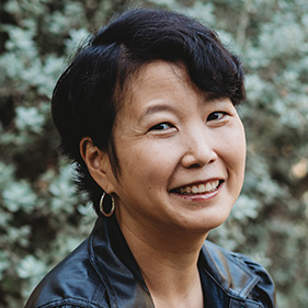 A smiling woman with dark hair and wearing dark jacket is seen against a background of green foliage. 