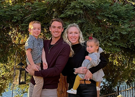 A man and a woman stand in a backyard under a tree, each holding one of their young children.