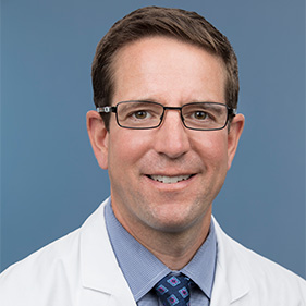 Man with light brown hair wearing a grey suit smiling at camera.