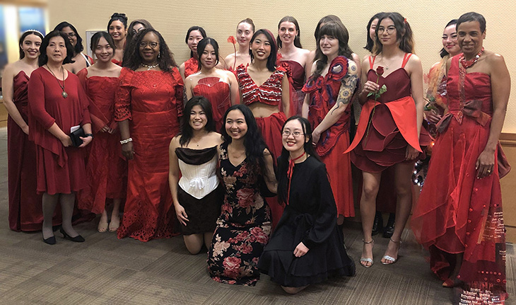 Two rows of women standing and wearing red dresses with three women wearing black and red kneeling in front of them.