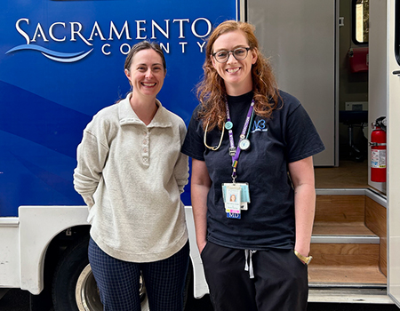 A woman in a beige sweater stands next to a woman in a blue shirt outside an RV with the words “Sacramento County” on the side