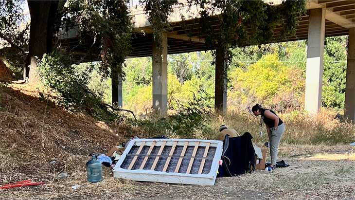 A woman stoops down to speak to a man in a wheelchair next to an abandoned mattress and debris near a homeless encampment 