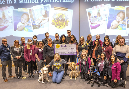 Twenty-four people and three dogs gather in a room holding an oversized check with $90,600 written on it