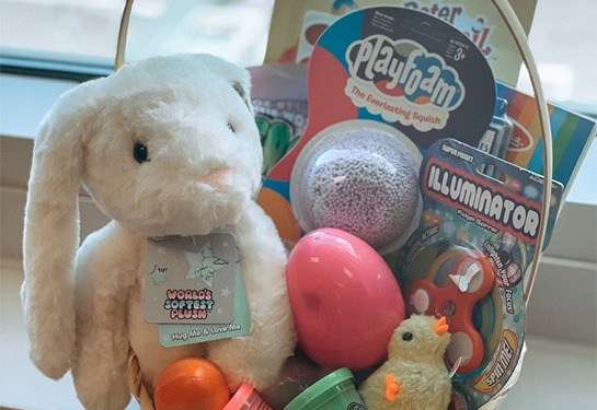 Basket on a windowsill is filled with a white stuffed rabbit, containers of slime, plastic eggs and a small stuffed chick. 