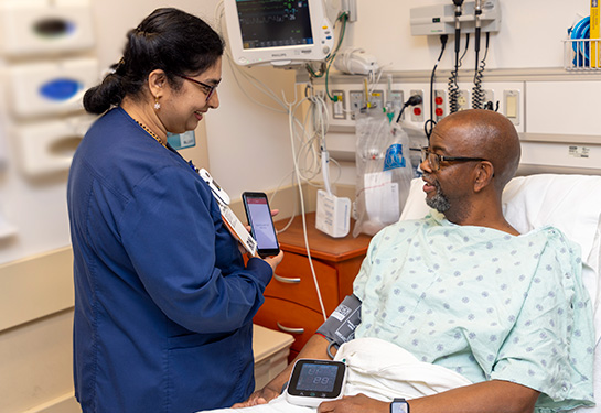 Woman in blue medical scrubs holds tablet while looking at patient lying in hospital bed.