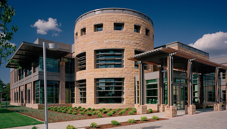 A large sandstone and glass building flanked by bushes and trees. 