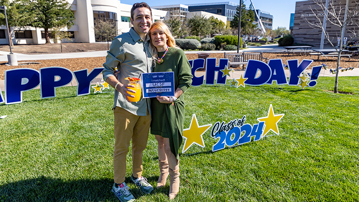 A man and a woman stand on grass at the medical school holding placard that states, "I matched UCSF Neurology"