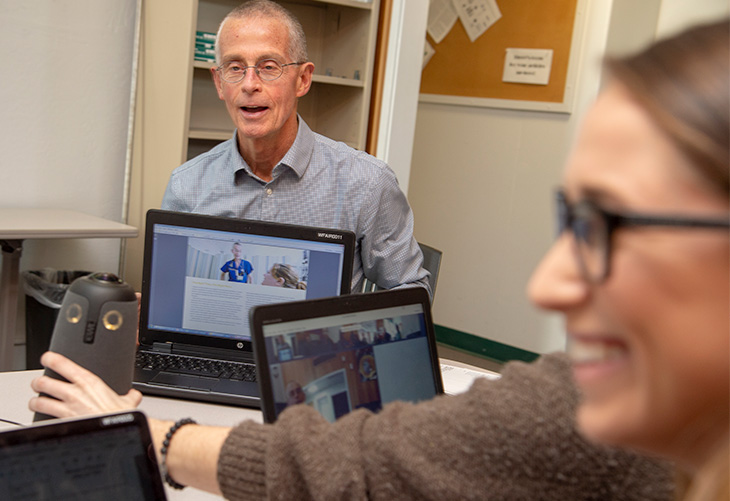 A man sits at a conference table and shows his laptop screen to others seated nearby