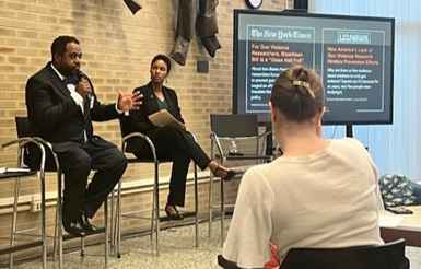 A seated man is talking into a microphone while a woman next to him watches. 