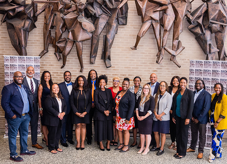 A group of 18 people stand together in front of a beige brick wall with a metal sculpture.