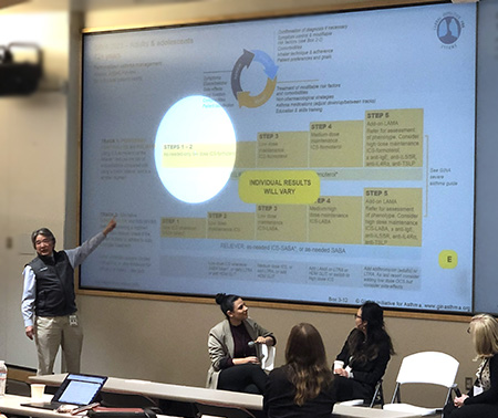 Man standing front of auditorium pointing at PowerPoint presentation on board.