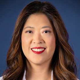 Alt Text for headshot: A surgeon with long brown hair, waring a white medical coat, smiles for a portrait.