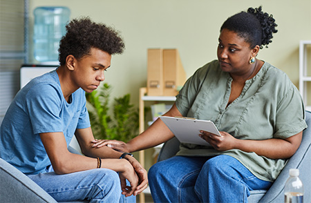 Young man wearing a blue t-shirt and blue jeans. He is looking down and leaning forward. A psychologist holding a folder in one hand and placing her other hand on the left elbow of the man. Both individuals have dark skin complexion.