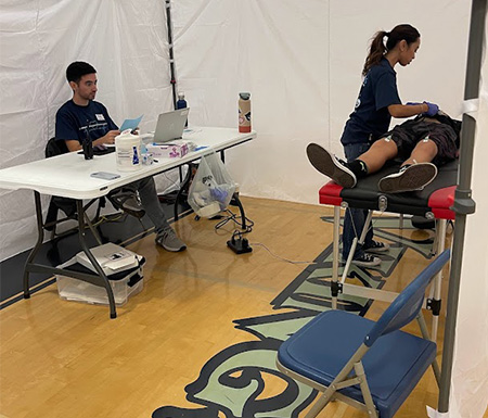 Inside of a medical tent, a boy lies on a table while a female volunteer puts probes on his chest. A man sits with a laptop at a table.