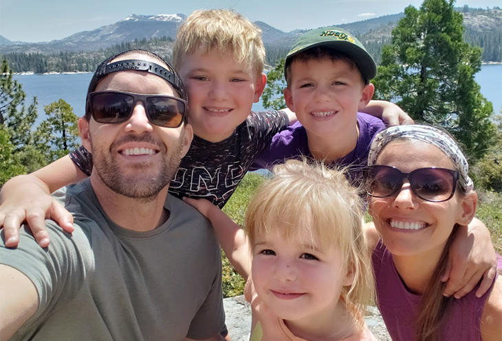 Man in sunglasses and woman in sunglasses and head scarf huddle with two young boys and a toddler girl with lake in background