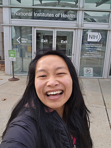 A woman with dark hair past her shoulders smiles in front of a glass doorway with the words “National Institutes of Health” on top