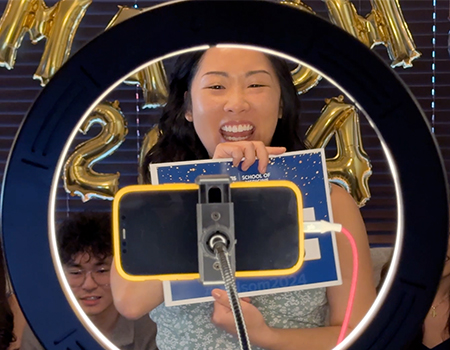 Young woman in blue dress smiles big looking into a phone surrounded by a ring light