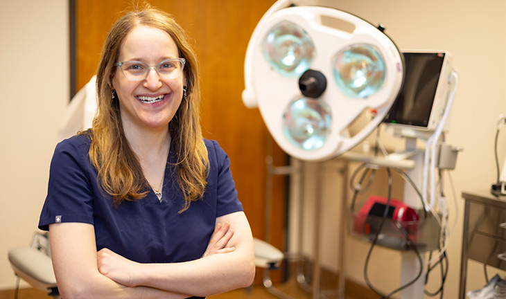 A woman with brown hair, wearing blue medical scrubs and glasses smiles in a dermatology office