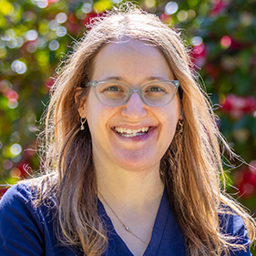 A woman with brown hair and glasses wearing navy blue medical scrubs stands outside near flowering trees. 