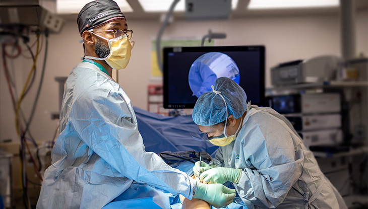 Two physicians in scrubs, masks and gloves operate on a patient’s knee. 