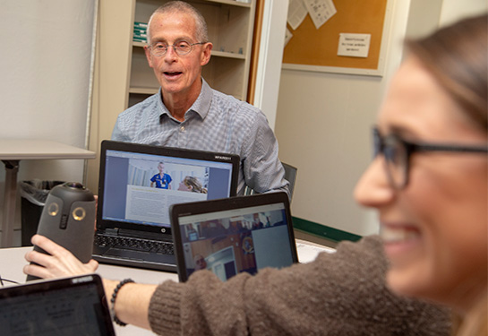 A man sits at a conference table and shows his laptop screen to others seated nearby