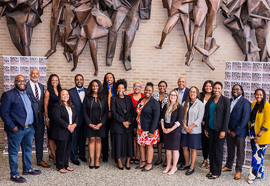 A group of 18 people stand together in front of a beige brick wall with a metal sculpture.