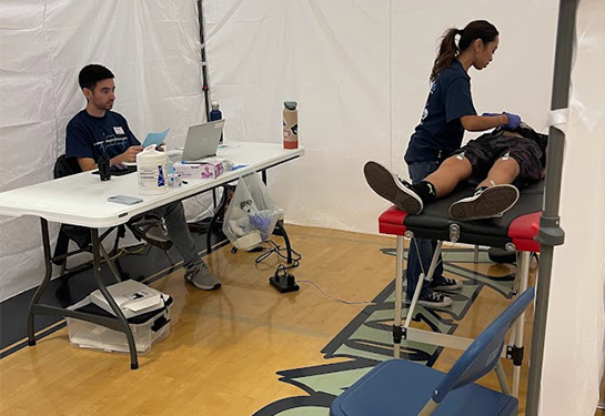 Inside of a medical tent, a boy lies on a table while a female volunteer puts probes on his chest. A man sits with a laptop at a table.