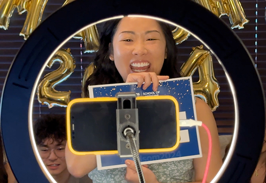 Young woman in blue dress smiles big looking into a phone surrounded by a ring light