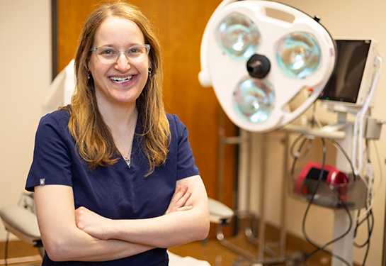 A woman with brown hair, wearing blue medical scrubs and glasses smiles in a dermatology office