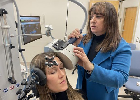 A woman with a black tracker on her forehead sits while another woman maneuvers a beige magnet coil near hear head.