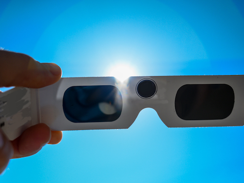 A hand holds a pair of eclipse glasses up toward the sun.