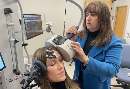 A woman with a black tracker on her forehead sits while another woman maneuvers a beige magnet coil near hear head.