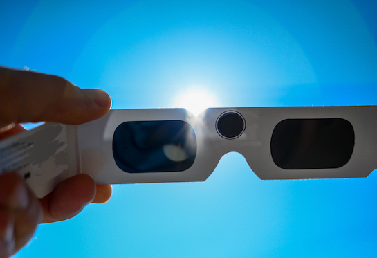 A hand holds a pair of eclipse glasses up toward the sun.