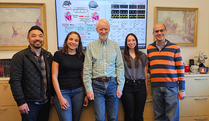 A group of three men and two women stand in front of a monitor with graphical representations of a scientific study.