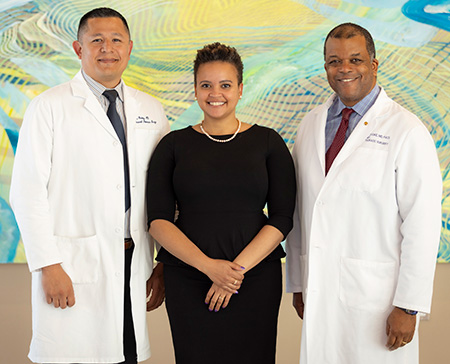 Man in white coat facing talking with woman in black dress and another man in white coat.