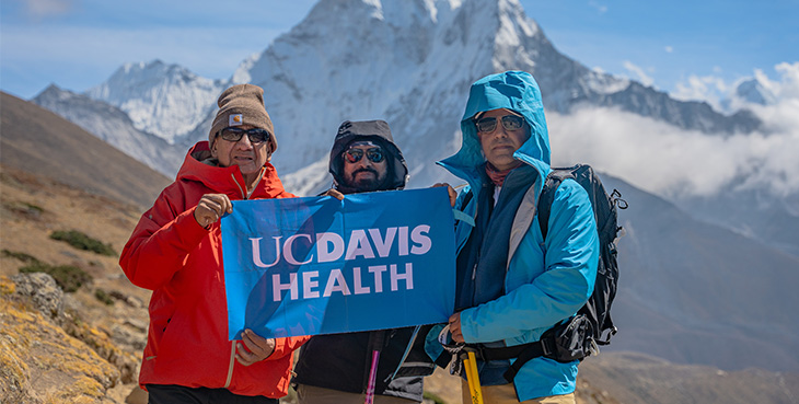 Tres hombres con chaquetas gruesas y gorras al frente al Monte Everest sosteniendo una bandea azul que dice: “UC Davis Health”