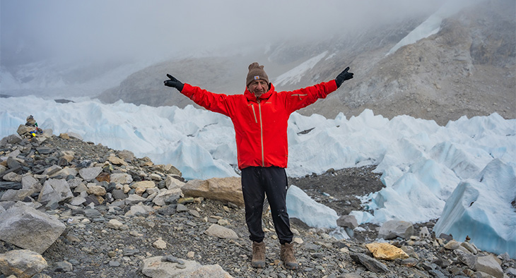 Un hombre con chaqueta roja, guantes negros y gorra color café levanta sus brazos en celebración al frente de una montaña nevada