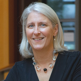 A woman with blond hair, wearing a black shirt and chunky necklace smiles. 