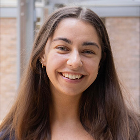 A woman with long brown hair, wearing a blue shirt, smiles outside while standing next to a brick building.