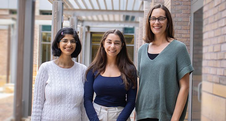 Three women of varying heights stand outside in a courtyard next to a brick building. 