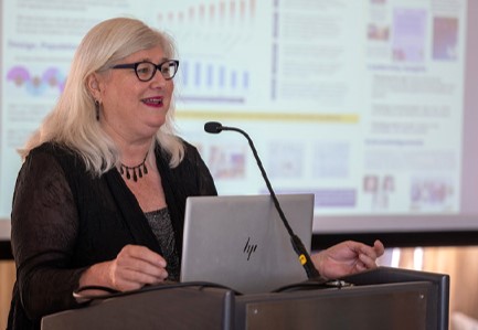 Woman speaks into mic at podium with PowerPoint screen behind her